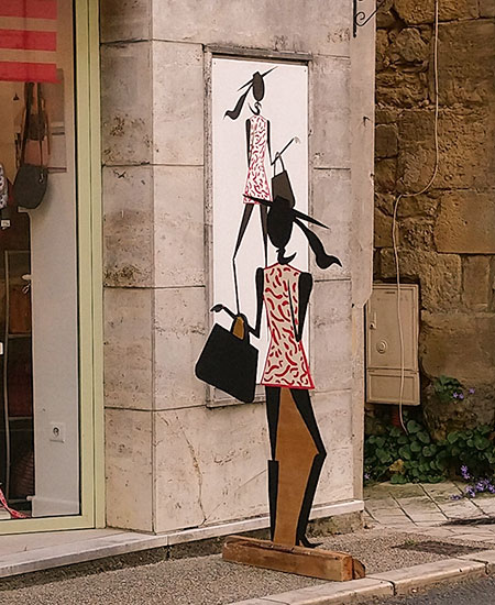 Panneau signalétique commerce (femme portant un foulard, un sac à mains et un chapeau peinte sur du bois) : vue de loin sur le trottoir